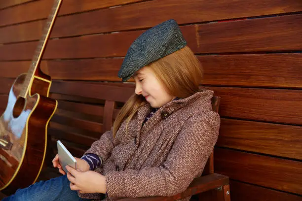 blond kid girl playing smartphone winter beret and guitar on wooden background