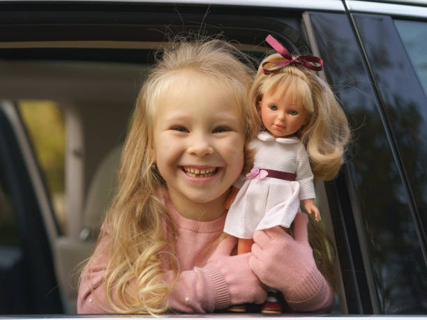 Portrait of happy girl with lovely toy doll Portriat of funny cute girl in the private car. She is sitting inside of  car, looking out of the car and holding a doll in her hands. Sunny autumn day. Concept of happy childhood girl playing with doll stock pictures, royalty-free photos & images