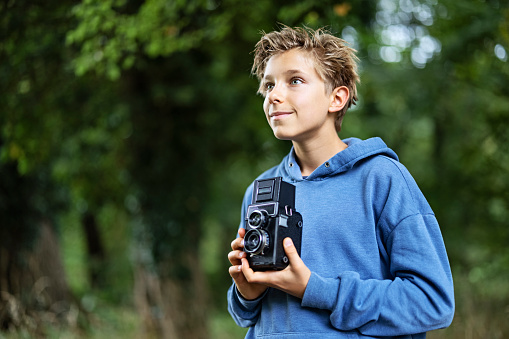 Smiling teenage boy is holding a vintage Twin-lens reflex camera camera. The boy is walking in the forest and taking photos.\nCanon R5