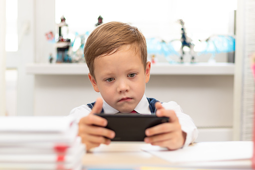 A cute first-grader boy in a school uniform on a light background at home sits at a desk with a smartphone in his hands. Selective focus. Close-up