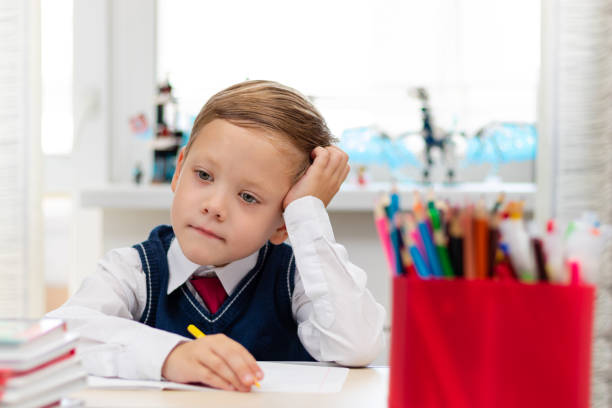 A cute boy first grader in a school uniform does homework while sitting at a desk with a pencil in his hands at home. Remote education. Selective focus. Close-up