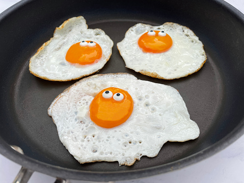 Stock photo showing close-up, elevated view of sunny side up fried eggs with googly eye cartoon faces cracked into a non-stick frying pan to be fried.