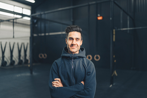 Smiling male athlete standing in gym. Sportsman is with arms crossed. He is wearing hooded shirt.