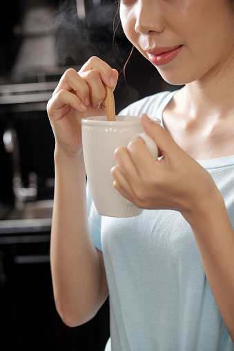 Close-up of content Asian girl stirrign sugar with spoon and smelling coffee scent in kitchen