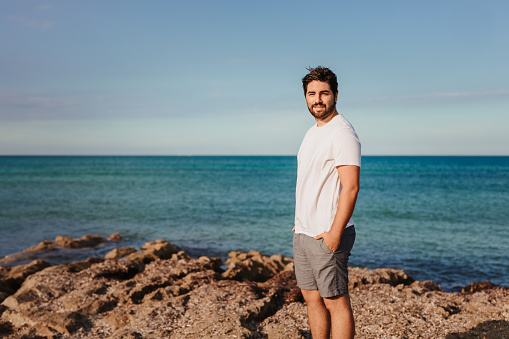 Handsome young man standing smiling at the camera in front of the beach. Useful for healthy, mindfulness, consciousness, awareness and self love concepts. Very selective focus and added grain. Part of a series.