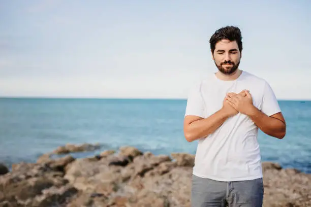 Handsome young man with hands on chest, closed eyes and grateful face gesture in front of the sea. Useful for healthy, mindfulness, consciousness, awareness and self love concepts. Very selective focus and added grain. Part of a series.