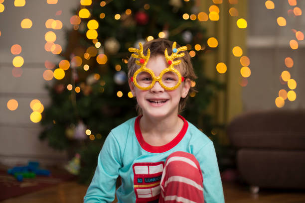 Cute little kid boy with party glasses and glowing Christmas garlands near a tree in home stock photo