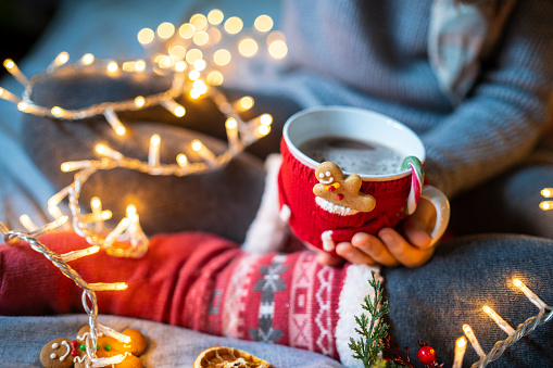 A woman sitting with legs up and wearing knitted Christmas stockings is holding a mug that is covered in a mini red sweater and a tiny gingerbread man in the jacket pocket, surrounded by illuminated Christmas lights