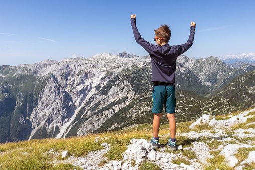 School-aged boy standing at the top of the hill, and looking at mountain tops, raising arms with fist pump gesture, celebrating mountaineering success