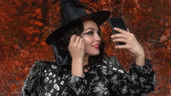 Portrait of a smiling girl in a witch hat and cloak. A girl in a carnival costume is drawn a face for Halloween in the open air. Outdoors. Face painting, headshot close up