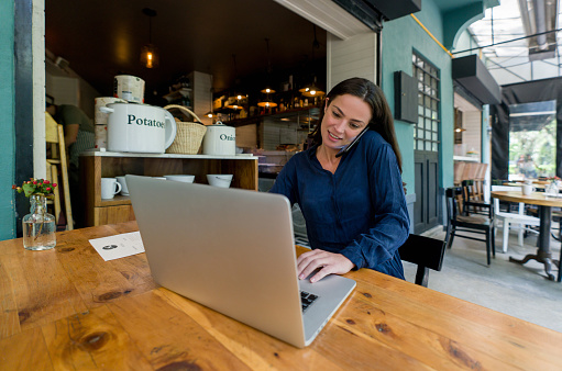 Latin American female entrepreneur working at a coffee shop on her laptop while talking on the phone