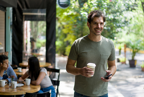 Happy man buying a cup of coffee to go at a cafe holding his cell phone on the other hand while smiling - lifestyle concepts