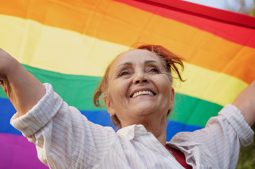 Beautiful mature woman with charming smile holding rainbow LGBT flag in her hands, gay and lesbian rights concept for all ages