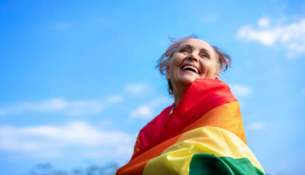 Beautiful mature woman with a charming smile holding a rainbow LGBT flag in her hands, gay and lesbian rights on a blue sky background stock photo