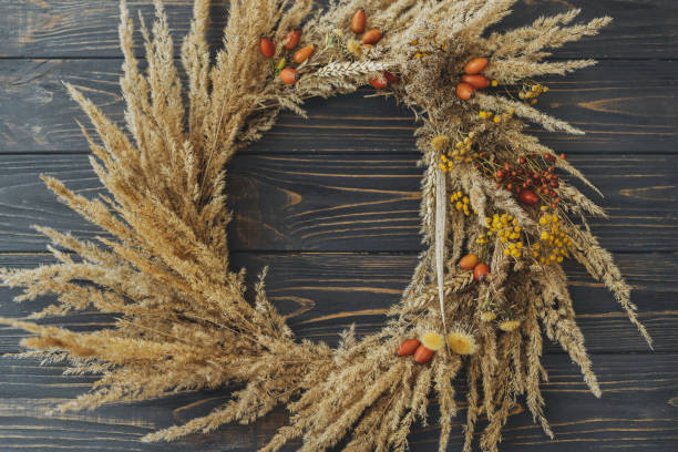 istock Stylish autumn rustic wreath flat lay. Creative boho wreath with dry grass, tansy wildflowers, wheat and dog-rose berries on rustic dark wooden table, top view. Holiday workshop 1344995101
