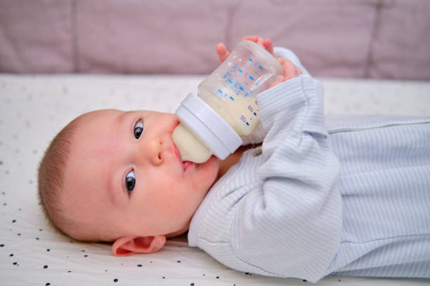 Happy baby is smiling on her own holding a bottle of milk in her hands. Three month old child keeps the mixture himself lying in bed Happy baby is smiling on her own holding a bottle of milk in her hands. Three month old child keeps the mixture himself lying in bed formula stock pictures, royalty-free photos & images