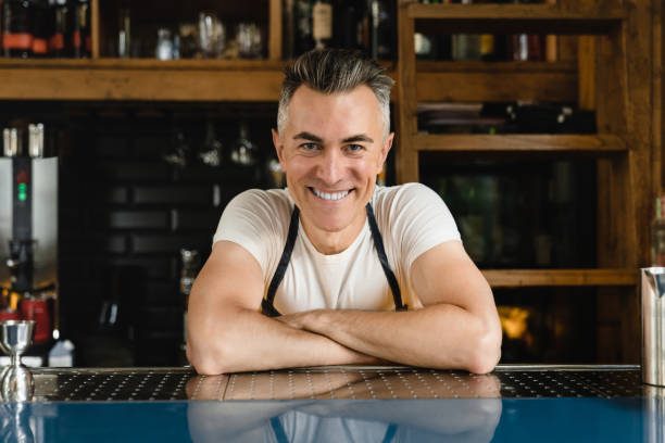улыбается зрелый кавказский бармен бариста владелец малого бизнеса официант в синем фартуке, опирающийся на барную стойку в ресторанном к� - manager bar counter restaurant wine стоковые фото и изображения