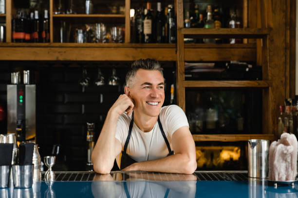 мечтательный улыбающийся кавказский зрелый бармен владелец малого бизнеса бариста официант опирается на барную стойку в синем фартуке в р - manager bar counter restaurant wine стоковые фото и изображения