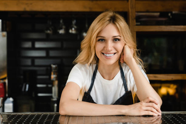 smiling caucasian young waitress small business owner barista bartender in blue apron looking at the camera standing at the bar restaurant counter - espresso women cup drink imagens e fotografias de stock