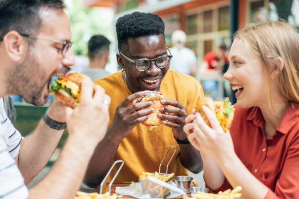 Friends and fast food Friends eating burgers and fries and have fun in outdoor restaurant mouth full stock pictures, royalty-free photos & images