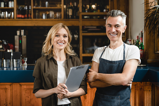 Smiling caucasian small business owner waiter barista with female auditor holding clipboard standing at the bar restaurant cafe counter checking products quality looking at the camera