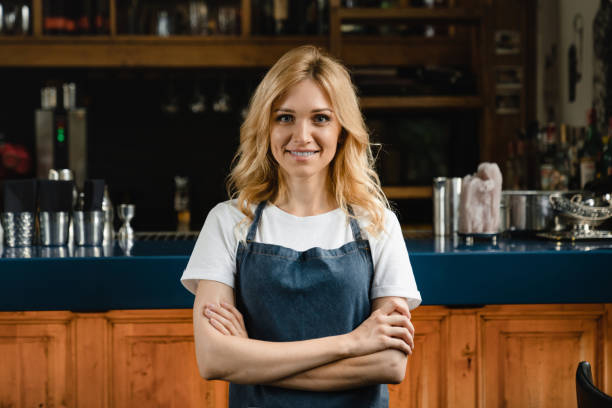 beautiful caucasian waitress small business owner barista bartender standing in blue apron looking at the camera with arms crossed at the bar counter in restaurant. - espresso women cup drink imagens e fotografias de stock