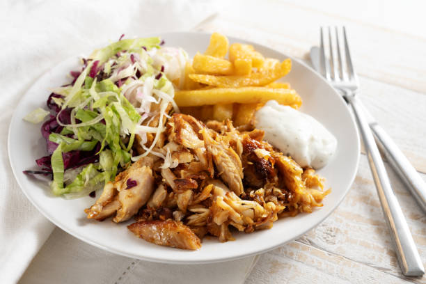 Doner kebab chicken meat with french fries, salad and tzatziki dip on a white plate, cutlery and napkin on a rustic wooden table, selected focus stock photo