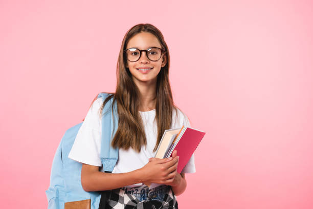 sorridente attiva eccellente studentessa studentessa in possesso di libri e libri di copia che vanno a scuola indossando occhiali e borsa isolati in sfondo rosa - school age girl foto e immagini stock