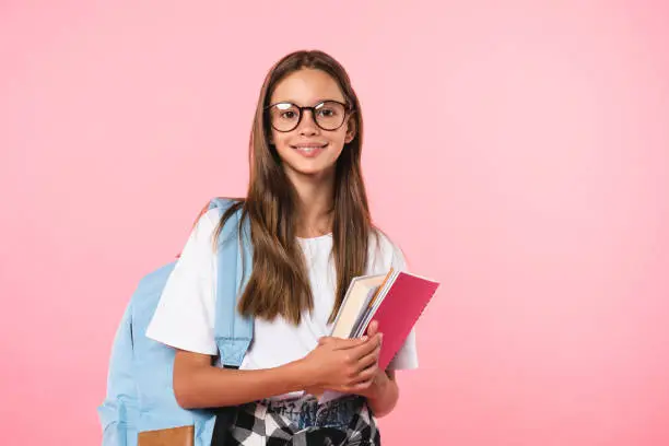 Photo of Smiling active excellent best student schoolgirl holding books and copybooks going to school wearing glasses and bag isolated in pink background