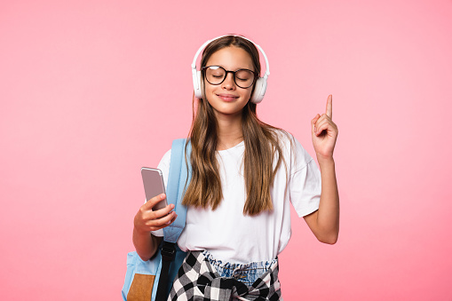 Happy caucasian schoolgirl teenager listening to the music going back to school pointing showing copy space isolated in pink background