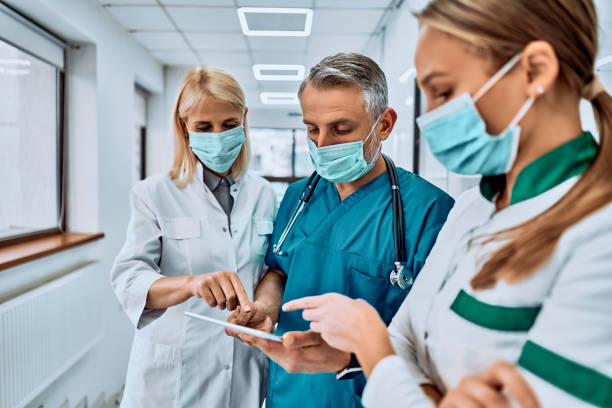 tre medici in maschera protettiva con un tablet sul corridoio dell'ospedale che controllano qualcosa. - doctor healthcare and medicine human resources teamwork foto e immagini stock