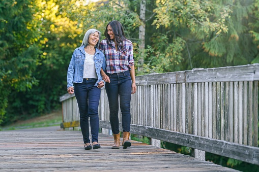 A vibrant senior woman of Asian descent links arms with her mixed race adult daughter as they walk across a footbridge in a natural parkland area. The family is enjoying time together outdoors while staying active and healthy.