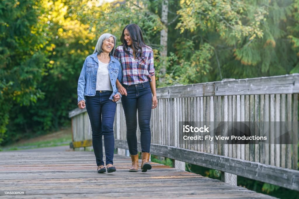 Senior Asian woman cherishing time with her adult daughter A vibrant senior woman of Asian descent links arms with her mixed race adult daughter as they walk across a footbridge in a natural parkland area. The family is enjoying time together outdoors while staying active and healthy. Mother Stock Photo