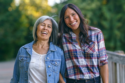 A vibrant senior woman and her adult daughter link arms affectionately and laugh while out on a an evening walk. The women are of Asian and Pacific Islander descent.