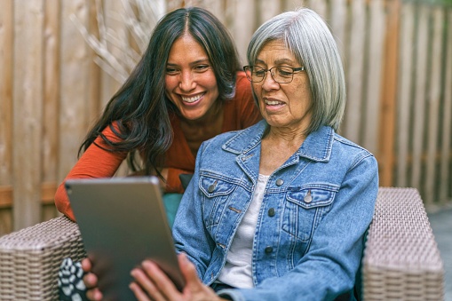 A senior woman of Asian descent and her adult daughter use a tablet computer to video call loved ones while social distancing due to the Covid-19 pandemic. The women are outdoors at home on the back patio. The older woman is sitting on outdoor furniture and her daughter is crouching down next to her. They are smiling and looking at the tablet computer.