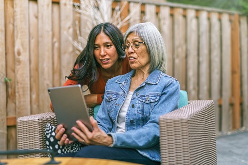 A retired mixed race senior woman sits on outdoor furniture on the back patio of her home. She is holding a tablet computer and her adult daughter is teaching her how to use it.