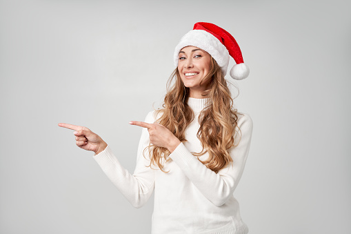 Woman christmas Sants Hat sweater white studio background Beautiful caucasian female curly hair portrait Happy person positive emotion Holiday concept Shows finger on side cope space for advertise