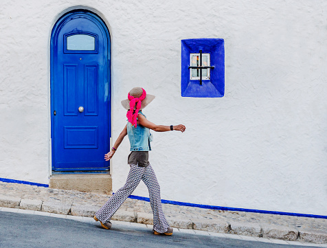 a caucasian woman walks quickly down a street in front of a white facade with blue doors and windows. she stands out against the white background. summer concept and tourism in Sitges, Spain.