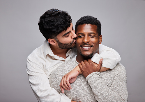 Young gay man hugging his smiling partner from behind and kissing him on the cheek against a gray background