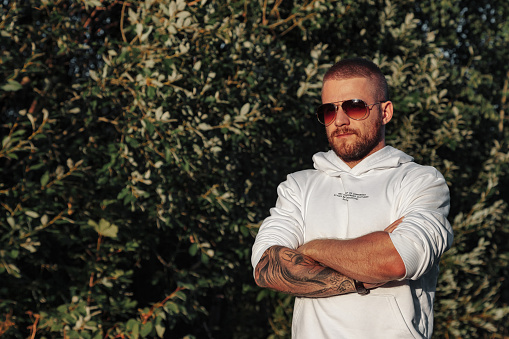 Portrait of young bearded man with tattoos in white jacket in countryside or in park. Male in casual clothes spends evening in nature. Concept of style, walking in fresh air and unity with nature