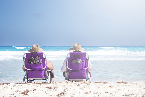 Two unrecognizable people in straw hats sit in chairs on caribbean beach, travel destinations