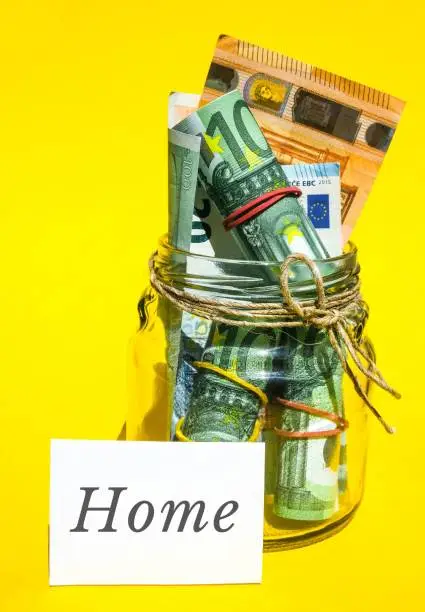 Photo of Glass jars filled with Euro bills, savings inside glass jar, money isolated on yellow background. Paper note written word HOME. Business budget of wealth