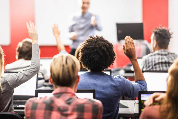 Back view of large group of students raising their arms during a class at computer lab. Focus is on man with curly hair.
