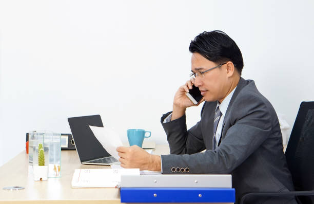 istock Asian businessman in suit talking on mobile phone while looking at report, personal working with laptop in the office. 1345080108