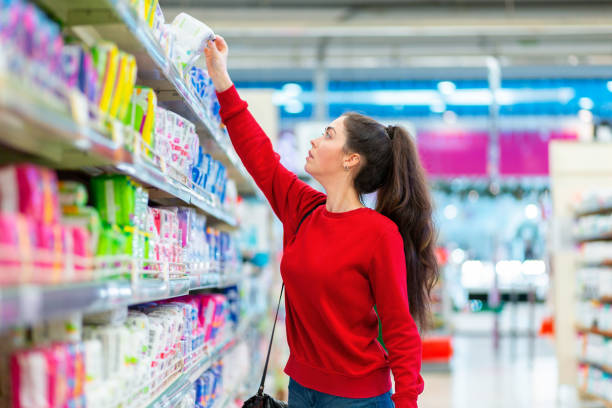 A young Caucasian woman reaches to the top shelf for a pack of sanitary pads. Side view. Corridor of shop on the background. The concept of purchasing hygiene products and cosmetics A young Caucasian woman reaches to the top shelf for a pack of sanitary pads. Side view. Corridor of shop on the background. The concept of purchasing hygiene products and cosmetics. sanitary napkin stock pictures, royalty-free photos & images