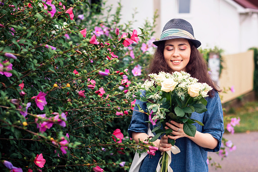 A smiling beautiful woman in a straw hat holds a bouquet of white roses and looks at him. Outside. The concept of March 8 and Mother's Day.