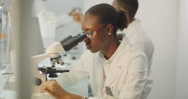 istock African, female scientist using microscope. Laboratory glassware behind her. Team in background 1345098543