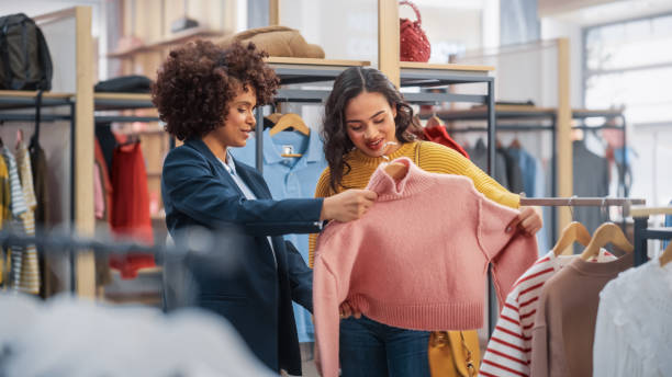 cliente joven comprando en tienda de ropa, asociado de ventas minoristas ayuda con asesoramiento. personas diversas en la tienda de moda, eligiendo ropa elegante, marca colorida con diseños sostenibles - shopping clothing store women retail fotografías e imágenes de stock