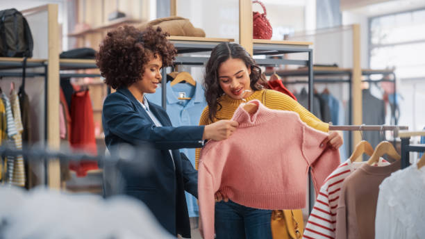 의류 매장에서 젊은 여성 고객 쇼핑, 소매 판매 동료는 조언에 도움이됩니다. 세련된 상점에서 다양한 사람들, 세련된 옷을 선택, 지속 가능한 디자인다채로운 브랜드 - 상점 이미지 뉴스 사진 이미지