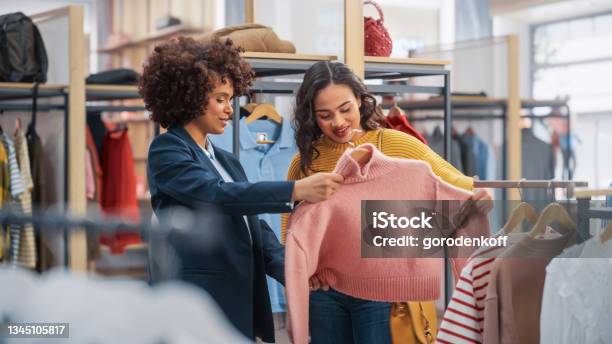 Young Female Customer Shopping In Clothing Store Retail Sales Associate Helps With Advice Diverse People In Fashionable Shop Choosing Stylish Clothes Colorful Brand With Sustainable Designs Stock Photo - Download Image Now
